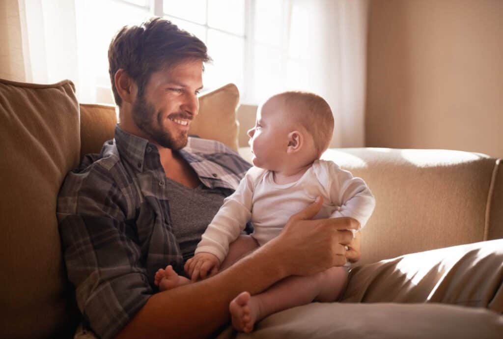 Vater hält Baby liebevoll auf dem Sofa, gemütliches Zuhause, natürliche Beleuchtung.