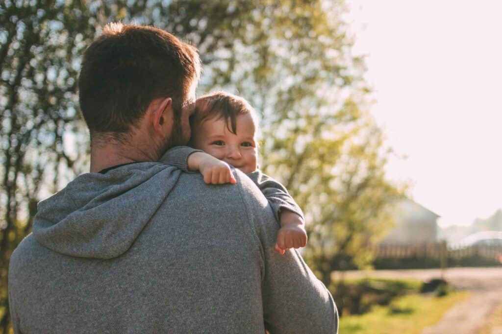 Vater hält lachendes Baby im Freien bei Sonnenuntergang, Natur im Hintergrund.