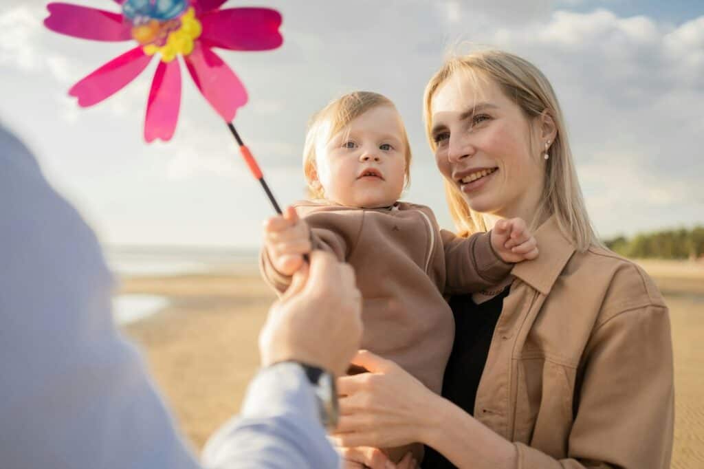 Mutter hält Baby am Strand, während jemand ein Windrad mit Blume zeigt.