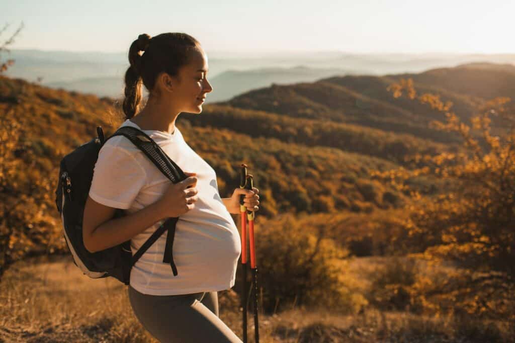 Schwangere Frau beim Wandern in der Natur mit Rucksack und Wanderstöcken.