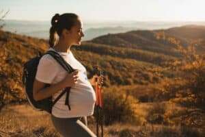 Schwangere Frau beim Wandern in der Natur mit Rucksack und Wanderstöcken.